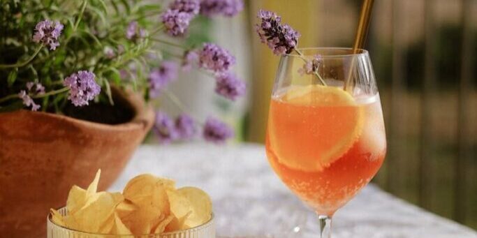 Outdoor Aperol spritz with chips on table, lavender background.